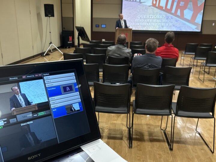 Conference room with a man speaking at a podium, viewed from behind a control monitor.